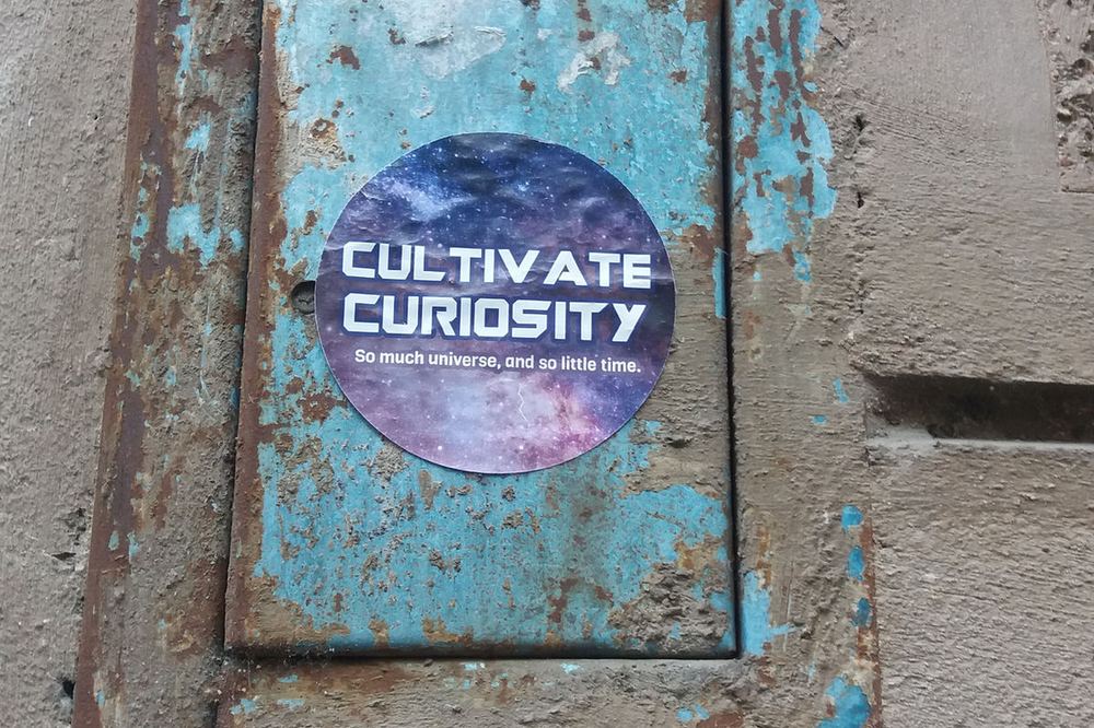 A sticker reading Cultivate Curiosity on a weathered blue wall